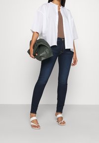 Camisa blanca de botones, blusa marrón, jeans oscuros y sandalias blancas. La modelo sostiene un bolso de cuero verde. Conjunto casual con texturas variadas.