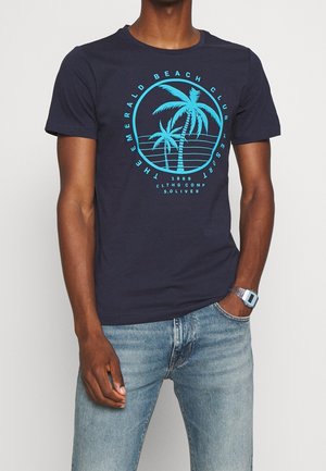 Man draagt een marineblauw T-shirt met een turquoise palmboomgrafiek en de tekst "Emerald Beach Club Resort", gecombineerd met lichtblauwe jeans en een zilveren horloge.