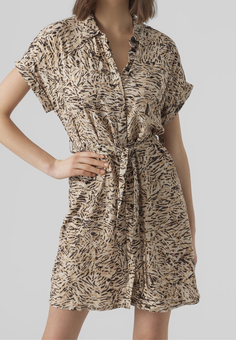 Femme portant une robe beige à manches courtes avec imprimé animal abstrait noir et marron, boutonnée devant et ceinture nouée à la taille.
