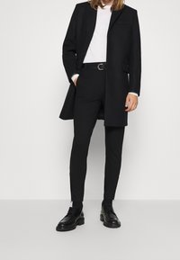 Abrigo de lana negro, jersey de cuello alto blanco, pantalones negros ajustados y zapatos negros con cordones y suela gruesa, acentuados con un cinturón.