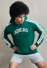Grön hoodie med vit Adidas-logga och tre vita ränder på ärmarna, tillsammans med grå shorts. Tyget verkar mjukt med en avslappnad passform.