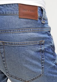 Jeans de mezclilla azul con un acabado desgastado, que presentan un parche de cuero marrón etiquetado "ONLY & SONS" y bolsillos traseros estándar con costuras de contraste.