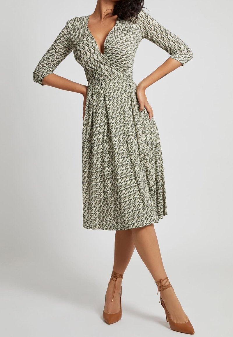 Femme portant une robe portefeuille mi-longue à motifs, manches trois-quarts, et des chaussures à talons hauts couleur beige, debout les mains sur les hanches.