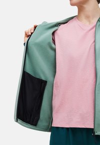 Persoon met een roze T-shirt houdt een lichtgroen jack open om de zwarte binnenzak met ritssluiting te laten zien.