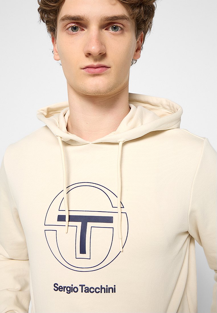 Haut à capuche beige en tissu doux avec un cordon. Présente un logo circulaire bleu marine et le texte "Sergio Tacchini" à l'avant.