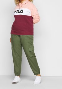 Bordeauxrode en roze hoodie met Fila-logo, gecombineerd met olijfgroene cargobroek met zijzakken en witte sneakers.