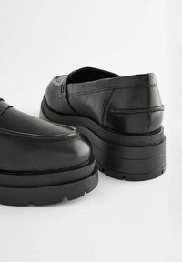 Mocassins en cuir noir avec une semelle épaisse et texturée. Tige lisse avec des détails de couture et bout arrondi. Design moderne et minimaliste.