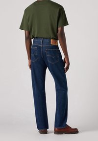 Homme portant un jean bleu foncé à coupe droite, un t-shirt vert olive et des chaussures en cuir marron, debout de dos sur un fond neutre.