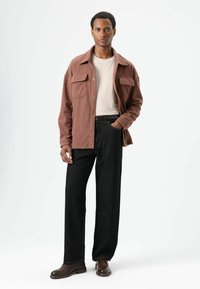 Bruine fleece overhemdjas met twee borstzakken, gecombineerd met zwarte rechtvallende jeans en bruine schoenen. Effen lichtgekleurd T-shirt eronder.