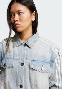 Casaco de ganga azul claro com dois bolsos no peito, botões pretos e três riscas brancas nas mangas. Apresenta um logótipo subtil no peito.