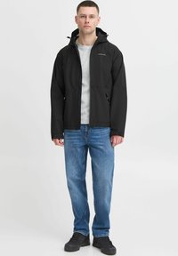 Črna kapuca z zadrgo na sprednji strani, oblečena preko sive srajce, v kombinaciji z modrimi džinsi in črnimi čevlji. Gladek, vodoodporen material.