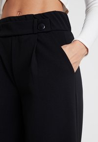 Pantalon noir avec une taille haute, doté d'une fermeture boutonnée à l'avant et de poches latérales. Le tissu est lisse avec une coupe ajustée.