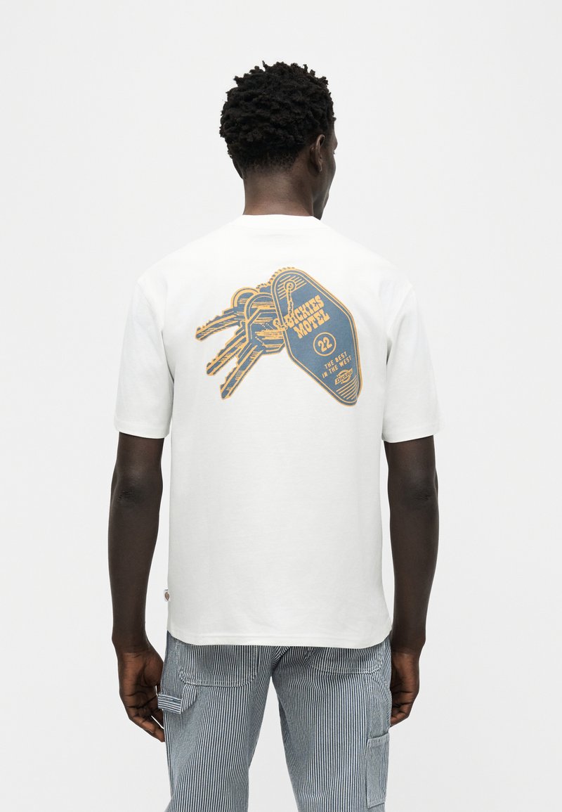 Man wearing white t-shirt with graphic of motel keychain labeled "Crickets Motel 22, The Best in the West" on back, striped pants.