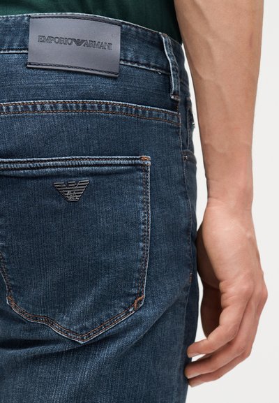 Κοντινό πλάνο πίσω τσέπης μπλε τζιν με το λογότυπο Emporio Armani και ετικέτα πάνω από τη μέση, δίπλα στο δεξί χέρι ενός ατόμου.