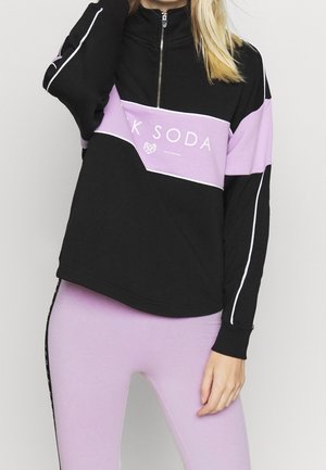 Vrouw draagt een zwart en lavendelkleurig sweatvest met rits en de tekst "Pink Soda" en bijpassende lavendelkleurige legging.