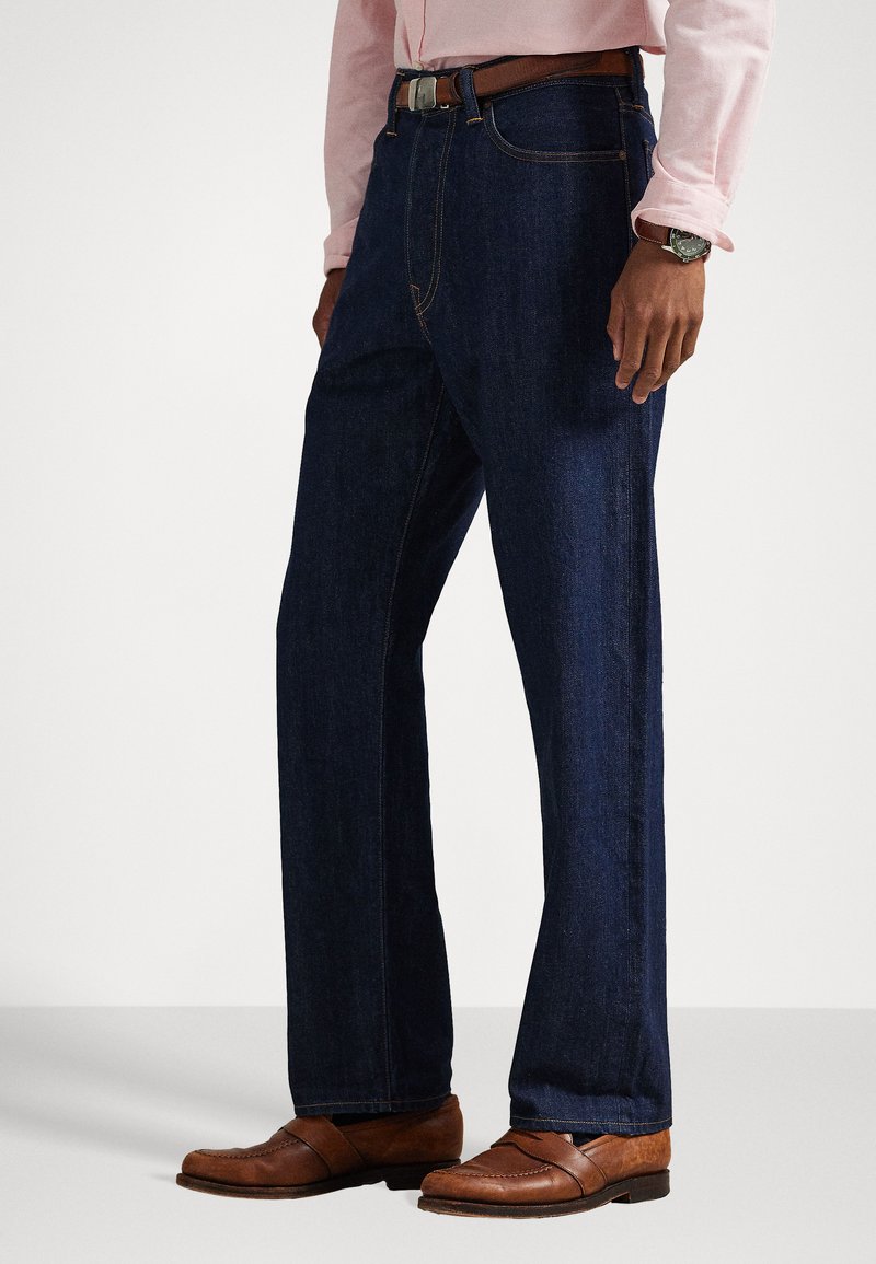 Dark blue denim jeans with a straight-leg cut, featuring contrasting stitching, a brown leather belt, and brown dress shoes.