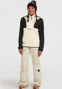 Crème en zwarte fleece pullover met een half-rits voorkant en logo, gecombineerd met crème sneeuwbroek en bruine laarzen. Zwarte beanie met pompon.