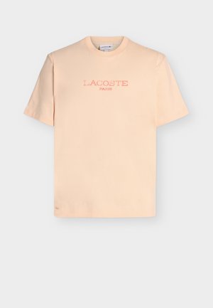 Ανοιχτόχρωμο ροδακινί κοντομάνικο μπλουζάκι με την επιγραφή "LACOSTE PARIS" σε ξεθωριασμένο ροζ μπροστά και μικρό κεντημένο κροκόδειλο κοντά στο τελείωμα.