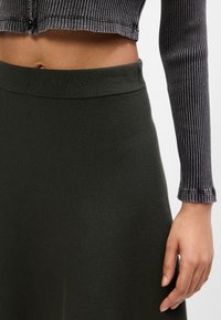 Žena v rebrovanom šedom dlhom rukáve crop top, ktorý má vysoký pás, a v tmavozelenej sukni, odhaľuje brucho a pravá ruka je uvoľnená pri boku.