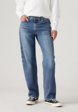 Personne portant un jean droit bleu clair, un sweat-shirt blanc et des baskets noir et blanc, debout devant un fond uni.