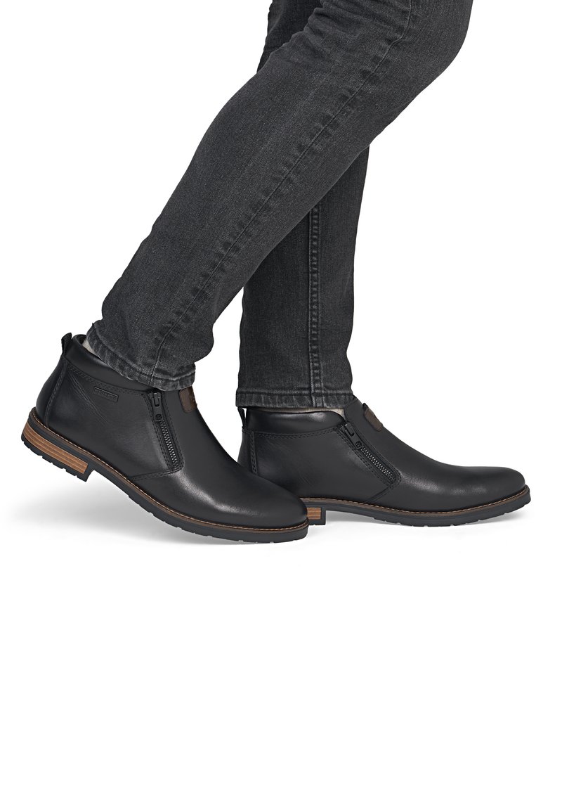 Personne portant un jean foncé et des bottines en cuir noir avec des fermetures éclair latérales et des semelles marron, debout les pieds croisés.
