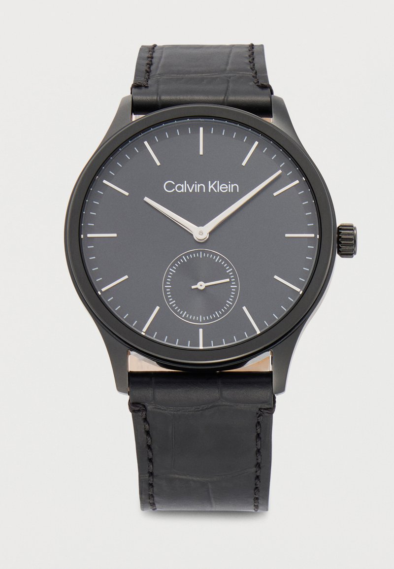 Μαύρο ρολόι χειρός Calvin Klein με μινιμαλιστικό καντράν, ασημένιους δείκτες ώρας και λεπτών, μικρό καντράν για τα δευτερόλεπτα και μαύρο δερμάτινο λουράκι.