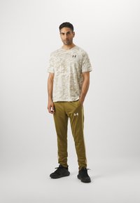 Camo-mønstret kortærmet skjorte i beige og olivengrønne sweatpants. Sorte atletiske sko; logo accenter på skjorte og bukser.