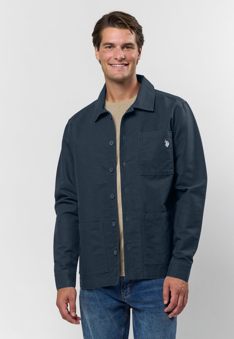 Homme souriant portant une veste bleu marine à boutons avec trois poches par-dessus une chemise beige et un jean bleu, debout devant un fond clair uni.