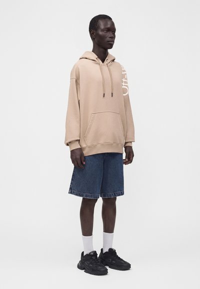 Personne portant un sweat à capuche beige avec un texte blanc sur la manche, un short en jean bleu à hauteur des genoux, des chaussettes blanches et des baskets noires, debout de côté.