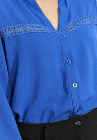 Blue button-up blouse with lace details across the chest, tucked into high-waisted black pants with a black button.