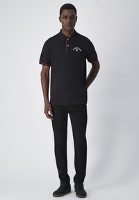 Zwarte poloshirt met rode accenten, voorzien van een ronde logo en een drieknoopsluiting. Gecombineerd met zwarte broek en gestructureerde sneakers.