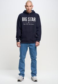 Jūrinės spalvos gladus su "BIG STAR" tekstu ir kišene, derinamas su šviesiai mėlynomis džinsomis ir pilkų sportinių batelių. Paprastas dizainas, kasdieniai audiniai.
