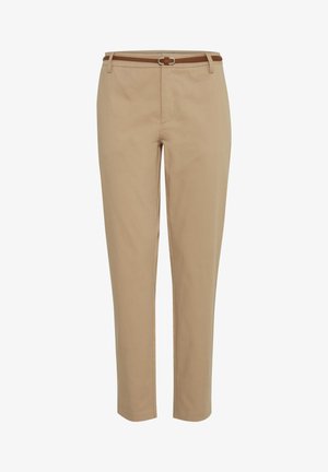 Frau trägt ein weißes T-Shirt, beige schlanke Hose mit Gürtel, weiße Turnschuhe, steht mit Händen in den Taschen vor einem einfarbigen Hintergrund.