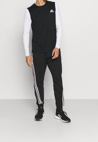 Uomo che indossa una maglietta senza maniche nera Adidas, una maglietta a maniche lunghe bianca come sottogiacca, pantaloni da allenamento neri Adidas con strisce bianche e scarpe da ginnastica nere Adidas.