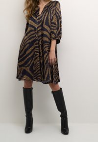 Lång svart klänning med ett djärvt brunt leopardmönster. Den har V-ringning, trekvartsärmar och en utsvängd silhuett, parat med knähöga svarta stövlar.