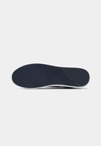 Semelle de chaussure en caoutchouc avec des rainures diagonales et horizontales, affichant la marque "Tommy Hilfiger" sur fond blanc.