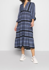 Robe longue à motifs avec des dessins géométriques bleus et noirs, un col en V et des manches amples. Portée avec des baskets blanches.