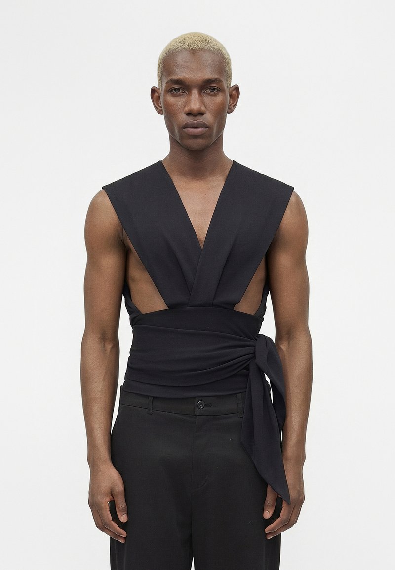 Λεπτό άτομο με κοντά ξανθά μαλλιά που φοράει μαύρο αμάνικο τοπ τύπου wrap με βαθύ V-λαιμό και μαύρο παντελόνι, στέκεται μπροστά σε απλό φόντο.