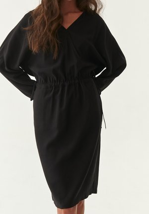 Frau trägt ein lockeres schwarzes Langarmkleid mit geschnürter Taille und V-Ausschnitt, steht vor einem schlichten hellen Hintergrund.