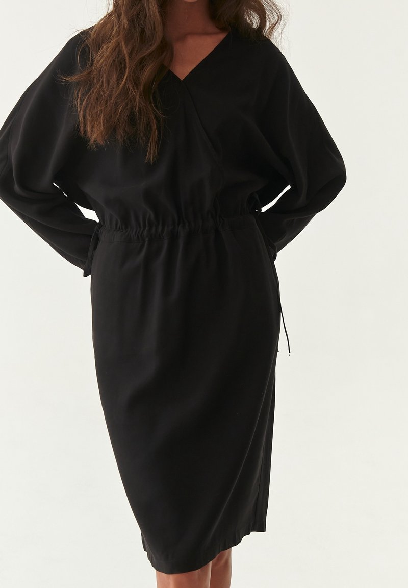 Femme portant une robe noire ample à manches longues avec une taille cintrée et un col en V, debout devant un fond clair uni.