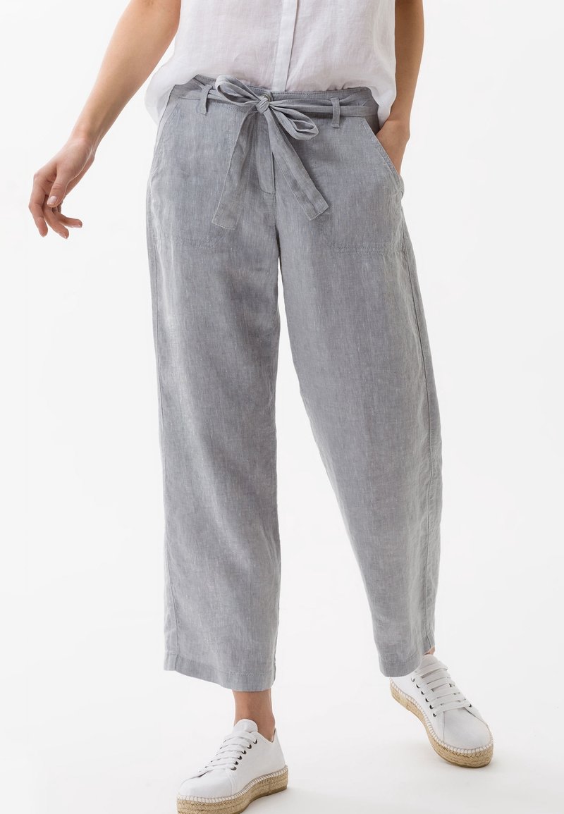 Femme portant un pantalon large gris clair avec une ceinture nouée à la taille et des baskets blanches, debout devant un fond blanc.