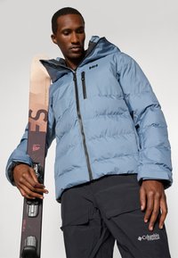 Svetlomodrá puffová bunda s kapucňou, s čiernymi zipsami a detailmi. Tmavé cargo nohavice s logom Columbia Titanium. Lyža v ruke.
