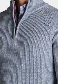 Gebreide pullover in lichtblauw met een textuur in ribbelpatroon, met een halve ritssluiting en rode accenten, en een gelaagde kraag over een shirt.