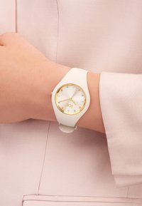 Orologio in silicone bianco con cassa dorata, quadrante argentato e indicatori delle ore colorati. L'orologio è indossato su un polso con uno sfondo in tessuto rosa chiaro.