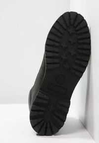 Semelle noire de bottine Timberland montrant des rainures profondes et un logo, inclinée contre un fond blanc de sol et mur.