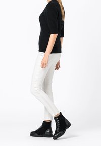 Maglione nero a maniche corte, pantaloni bianchi slim-fit e stivaletti neri alla caviglia con lacci e dettagli testurizzati. L'outfit presenta un look moderno e contrastante.