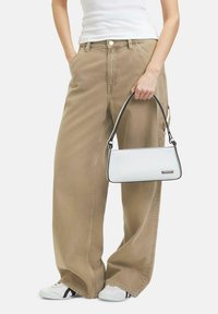 Borsa bianca con dettagli neri, forma rettangolare slim, tenuta in mano. Indossata con pantaloni beige a gamba larga e sneaker bianche.