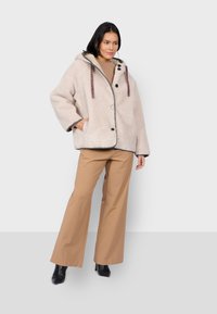 Beżowa puszysta kurtka z kapturem, zapinana na guziki, z wstążkami w panterkę. W połączeniu z beżowymi szerokimi spodniami i czarnymi botkami.