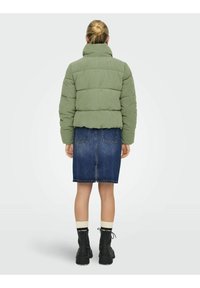 Chaqueta acolchada verde oliva con cuello alto, combinada con una falda vaquera. Botines negros con cordones y calcetines de color crema visibles.