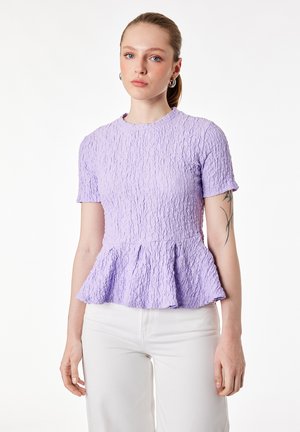 Młoda kobieta z jasnymi włosami związanymi w kucyk, ubrana w teksturowany lawendowy top z krótkim rękawem i baskinką oraz białe spodnie, stojąca na tle białego tła.